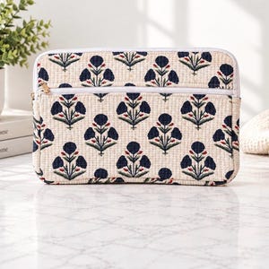 May include: A rectangular quilted laptop sleeve featuring an off-white base with a repeating dark blue floral pattern. The sleeve includes a horizontal zippered front pocket with a white zipper track and a white border trim along the edges.