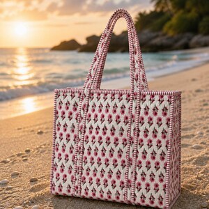 May include: A rectangular tote bag with a floral pattern in pink and white, set against a beach backdrop. The bag has matching pink handles and trim, and is sitting on the sand near the water.