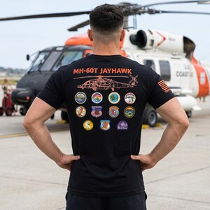 May include: A black short-sleeved t-shirt featuring an orange line drawing of an MH-60T Jayhawk helicopter on the back. Below the helicopter are eleven circular patches and emblems. The right sleeve displays a small orange American flag graphic.