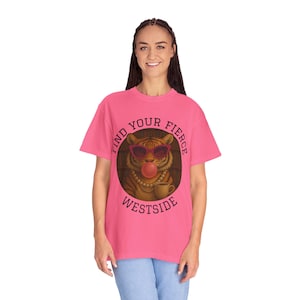 Pode incluir: Uma camiseta rosa com um gráfico de um tigre usando óculos de sol e soprando uma bolha de chiclete. O texto "FIND YOUR FIERCE WESTSIDE" é impresso ao redor da imagem do tigre. A camiseta está sendo usada por uma pessoa.