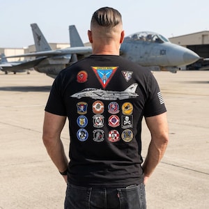 May include: A black short-sleeved t-shirt featuring a large graphic on the back. The design includes a central illustration of a fighter jet, a triangular Fighter Wing patch, and twelve circular military-style squadron patches. A small flag is on the sleeve.