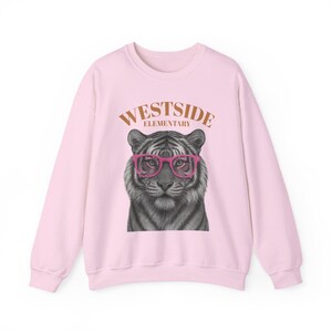 Pode incluir: Um moletom rosa claro com um tigre usando óculos rosa. O texto "WESTSIDE ELEMENTARY" está arqueado acima da cabeça do tigre em marrom. O moletom tem mangas compridas e gola redonda.