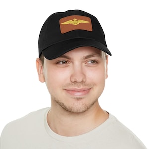 May include: Black baseball cap with a brown leather patch featuring a gold-colored emblem. The emblem has a wing design with a central crest. The cap is made of a soft material and has a curved brim.