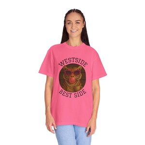 Pode incluir: Uma t-shirt rosa choque com o texto "WESTSIDE BEST SIDE" em torno de uma ilustração de um tigre a usar óculos de sol e a fazer uma bolha. O tigre também usa um colar de pérolas e segura uma chávena. A t-shirt é feita de um material macio.