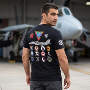 Op de afbeelding: Een zwart t-shirt met korte mouwen en een grote afbeelding op de achterkant. Het ontwerp bevat een centrale illustratie van een gevechtsvliegtuig, een driehoekige patch met de tekst Fighter Wing en talloze ronde squadronpatches. Een kleine Amerikaanse vlag bevindt zich op de mouw.