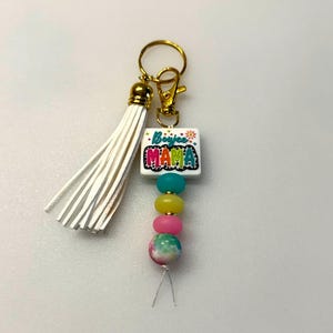 May include: A gold-toned keychain with a white tassel and a lobster clasp. The keychain features a white square with the words "Boujee Mama" in colorful letters, and a string of colorful beads in teal, yellow, pink, and a tie-dye pattern.