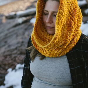 May include: A woman wearing a yellow crocheted cowl with a hood. The cowl is made with a textured stitch and has a gradient of color from a darker yellow to a lighter yellow.