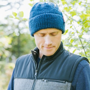 Puede incluir: Un hombre con un gorro azul de punto y un chaleco azul oscuro y gris. El chaleco tiene una cremallera y un bolsillo.