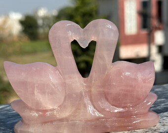 May include: A rose quartz sculpture depicting two swans with their necks intertwined to form a heart. The light pink stone sculpture sits on a rectangular base. The background shows a blurred view of greenery and a building. The sculpture is approximately 15 cm in length.