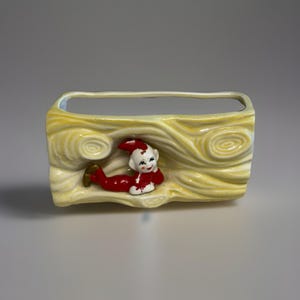Puede incluir: Una jardinera rectangular de cerámica amarilla con un diseño de vetas de madera texturizadas. Una pequeña figura de elfo con traje rojo, cara y gorro blancos está tumbada en la parte delantera. La jardinera tiene una abertura rectangular en la parte superior.