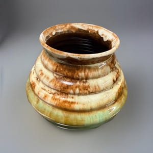 以下が含まれることがあります： 幅広で丸みを帯びたボディとわずかに広がった縁を持つ、手作りのセラミック花瓶。茶色、クリーム色、緑色などの水平な色の帯があります。内部は濃い茶色です。装飾品です。