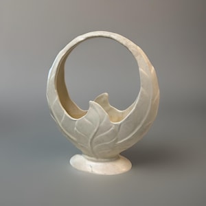 May include: Off-white ceramic basket with a handle. The basket features a leaf design and a rounded base. The handle is a full circle. The basket is a decorative item.