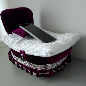 Może przedstawiać: Pluszowy, fioletowy aksamitny stojak na telefon z białą poduszką i małą fioletową poduszką. Stojak ma ozdobne srebrne wykończenie i czarne zapięcie na guzik. Czarny smartfon spoczywa na białej poduszce.