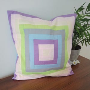 Może przedstawiać: Kwadratowa poduszka dekoracyjna z patchworkowym wzorem. Poduszka posiada koncentryczne kwadraty w pastelowych kolorach: lawendowym, jasnozielonym, niebieskim i szarym. Poduszka leży na drewnianej powierzchni.
