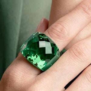 May include: A large, square-cut emerald green ring featuring a faceted surface that reflects light. The ring has a bold, chunky design with a polished finish, showcasing a deep, vibrant green color throughout the entire gemstone-like structure.