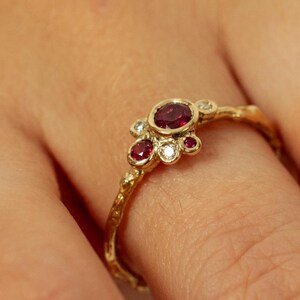 May include: A gold ring with a textured, organic-looking band featuring a cluster of three small red rubies and two clear white diamonds set in a bezel arrangement. The metal has a warm, yellow tone and a slightly uneven, natural surface finish.