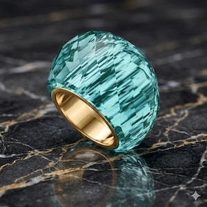 Könnte beinhalten: Ein facettierter, aquamarinblauer Edelsteinring mit einem goldenen Band. Die Oberfläche des Rings ist mit zahlreichen Facetten versehen, die einen funkelnden Effekt erzeugen. Das goldene Band sorgt für einen kontrastierenden warmen Ton. Der Ring liegt auf einer dunklen, marmorierten Oberfläche.