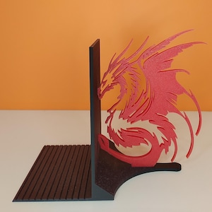 Può includere: Un fermalibri a forma di drago rosso e arancione. Il drago è tagliato da materiale a strati e presenta dettagli intricati. Il fermalibri è supportato da una base nera con una superficie strutturata. Lo sfondo è arancione con il testo "Teccà 3D SOLUTIONS".