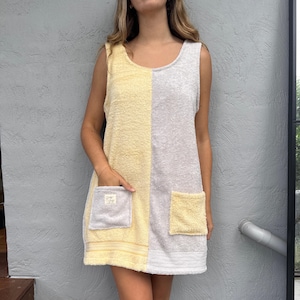 May include: A sleeveless terry cloth dress featuring a color-blocked design with a pale yellow left side and a light gray right side. The dress includes two contrasting patch pockets, one gray on the yellow side and one yellow on the gray side.