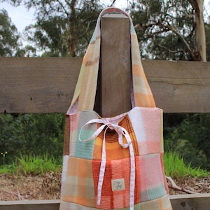 May include: A patchwork tote bag constructed from various plaid wool-like fabric squares in shades of orange, yellow, green, and white. The bag features a white ribbon drawstring closure with small pink dots and a rectangular label reading Shabby Vintage.