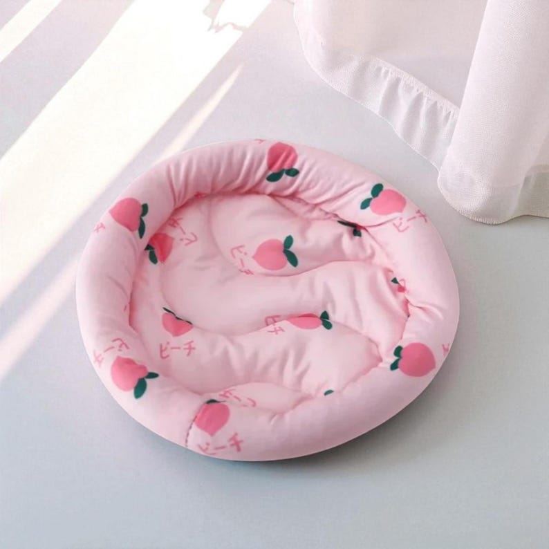 Puede incluir: Una cama redonda rosa para mascotas con un estampado de melocotones. La cama tiene un centro acolchado y un borde acolchado elevado. El dise&ntilde;o de melocotones incluye melocotones rosas con hojas verdes y texto japon&eacute;s.