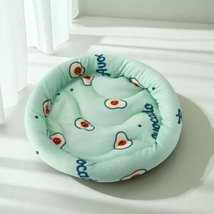 Puede incluir: Una cama redonda para mascotas de color verde claro con un patr&oacute;n repetido de aguacates. Los aguacates son blancos con centros rojos y est&aacute;n impresos sobre una superficie acolchada. La palabra "aguacate" est&aacute; impresa en azul alrededor del borde de la cama.