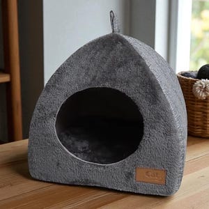 May include: A gray, enclosed cat bed with a round opening and a small handle on top. The bed is made of a soft, plush material and has a brown leather tag that reads "Cat". The cat bed is on a wooden surface.