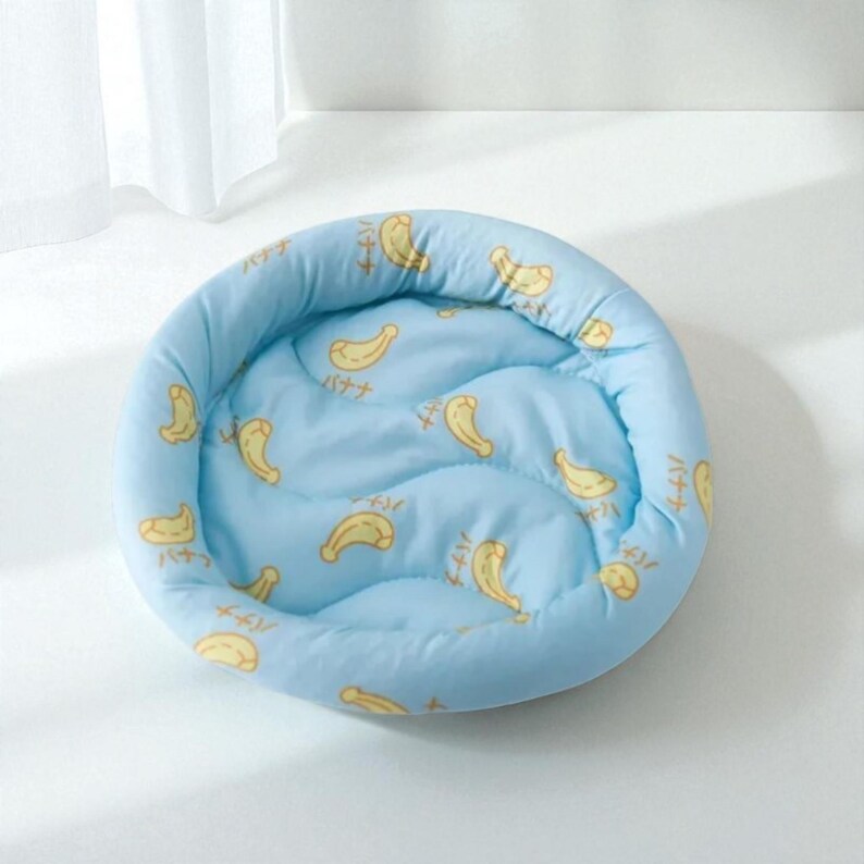 Puede incluir: Cama redonda para mascotas, de color azul claro, con estampado de pl&aacute;tanos amarillos y texto japon&eacute;s. La cama tiene un dise&ntilde;o acolchado para mayor comodidad. Adecuada para mascotas peque&ntilde;as, ofrece un espacio para dormir acogedor. Di&aacute;metro aproximado de 40 cm.