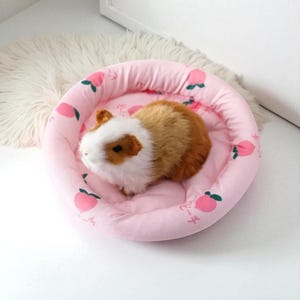 Puede incluir: Una cama rosa y redonda para mascotas con un estampado de melocotones y un conejillo de indias de peluche. La cama tiene un borde elevado y un interior suave y acolchado. El conejillo de indias tiene pelaje marr&oacute;n, blanco y canela.