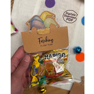 May include: A clear cellophane bag filled with candy, featuring a brown paper tag with the text "Fasching in der Tüte" and a jester hat illustration. The bag contains Haribo and Maoam candies.