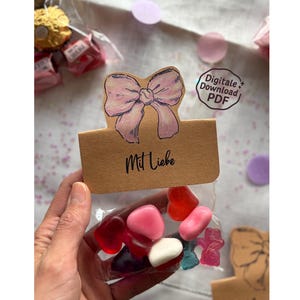 May include: A small cellophane bag filled with heart-shaped candies in various colors. The bag is attached to a brown card with a hand-drawn pink bow and the words "Mit Liebe". A digital download PDF sticker is visible.