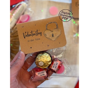 May include: A small gift bag with chocolates and a decorative tag. The tag is brown with a heart-shaped lock design and the text "Valentinstag in der Tüte". The bag contains a gold-wrapped chocolate and Mon Cheri candies.