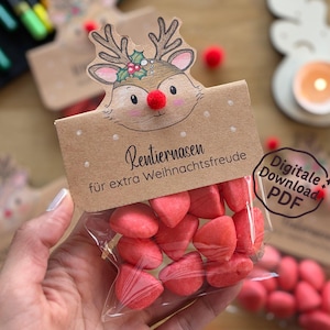 Puede incluir: Una bolsa transparente llena de caramelos rojos en forma de corazón, coronada con una etiqueta con temática de reno. La etiqueta presenta una ilustración de reno con una nariz de pompón rojo y el texto "Rentiernasen für extra Weihnachtsfreude."