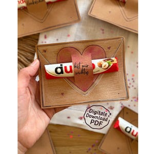 Può includere: Una busta di carta marrone con un motivo a cuore e una barretta di cioccolato Kinder. La barretta è avvolta in una confezione rossa e bianca. Il testo "du bist mir wichtig" è su un'etichetta a forma di cuore. L'immagine include anche il testo "Digitale Download PDF".