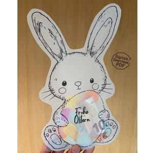 May include: A white bunny-shaped card with a black outline, holding a colorful egg. The egg has the text "Frohe Ostern" and the number "10" printed on it. The card is held against a wooden background.