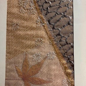 May include: A textile artwork with a leaf design. The artwork is divided diagonally, with a textured tan fabric on the left and a patterned brown mesh on the right. Gold trim and silver accents add detail.