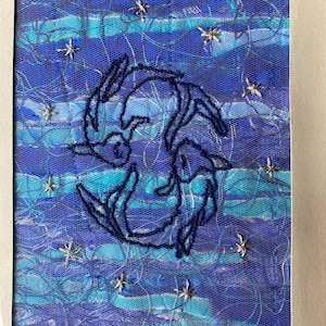 Puede incluir: Una obra de arte textil con un diseño bordado en azul oscuro de dos conejos entrelazados. El fondo es una mezcla de telas azules y turquesas con adornos de estrellas plateadas, todo enmarcado en blanco.