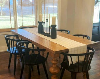 May include: A wooden dining table with a light-colored top and a turned leg base. Six black chairs surround the table, and a cream-colored table runner is centered on the table. Decorative candles and a chandelier are above the table.