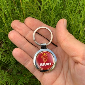 Könnte beinhalten: Ein silberner Schlüsselanhänger mit einem runden Anhänger, der das SAAB-Logo zeigt. Das Logo ist ein goldener Greif mit Krone auf rotem Grund, umgeben von einem silbernen Rand. Der Schlüsselanhänger wird in einer Hand vor grünem Laub gehalten.
