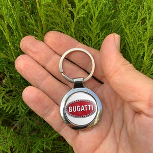 Puede incluir: Un llavero plateado con un colgante redondo con el logotipo de Bugatti. El logotipo es un óvalo rojo con la palabra "BUGATTI" en letras blancas, sobre un fondo blanco. El llavero se muestra sobre un fondo de follaje verde.