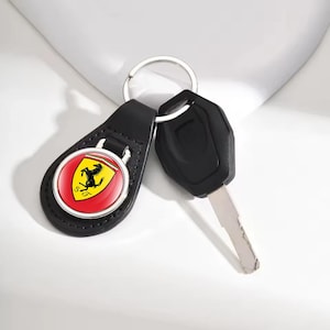 Könnte beinhalten: Schwarzer Leder-Schlüsselanhänger mit dem Ferrari-Logo in Rot, Gelb und Schwarz. Der Schlüsselanhänger ist an einem silbernen Schlüsselring befestigt, zusammen mit einem schwarzen Autoschlüssel.