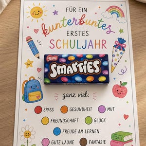 Puede incluir: Una tarjeta rectangular con el texto en alemán FÜR EIN kunterbuntes ERSTES SCHULJAHR y una caja de caramelos Nestle Smarties colocada en el centro. La tarjeta presenta ilustraciones coloridas de un sol, arcoíris, lápiz, mochila, manzana, libros y flores.