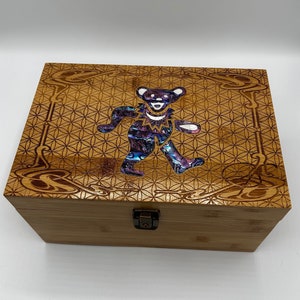 May include: A wooden box with a latch closure. The lid features a laser-etched design of a flower of life pattern and a colorful, iridescent bear.