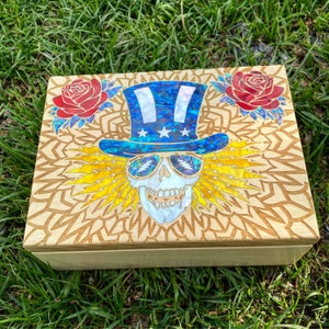 May include: A wooden box with a decorative design featuring a skull wearing a blue and white hat with stars. The skull has yellow lightning bolts around it and there are red roses on either side of the skull.