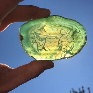 May include: A green agate slice with a geometric pattern and a hand-painted design of a person holding a turtle. The design is in a light yellow color.