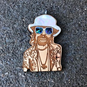 May include: A wooden ornament of a person with long hair wearing a white hat with iridescent accents, sunglasses, and a floral patterned shirt.