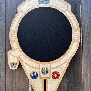 May include: Wooden Star Wars-themed wall art shaped like the Millennium Falcon. The art piece features a black circular center, light wood trim, and inlaid decorative elements. The Star Wars logo is at the top, and three symbols are at the bottom.