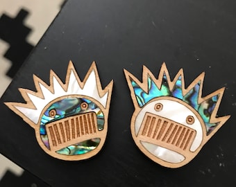 Puede incluir: Dos alfileres de madera con un diseño de concha iridiscente blanco y azul. Los alfileres tienen forma de cara con una corona de púas y una boca con una serie de líneas.