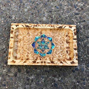May include: A wooden tray with a decorative floral design and a blue and green abalone shell inlay in the center. The inlay features a geometric star shape.
