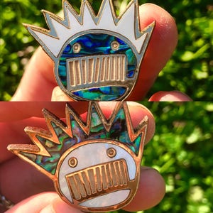 May include: Two enamel pins featuring a cartoon face with a crown of spikes. The first pin has a blue and green abalone shell design. The second pin has a white and gold abalone shell design.